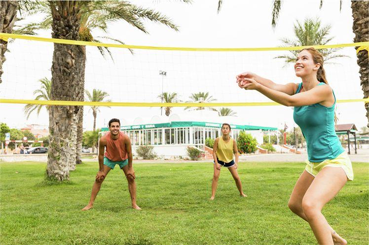 mladí lidé hrají v parku mezi palmami volejbal, dívka v zeleném tílku čeká na příjem míče, zpoza sítě ji sledují muž v oranžovém a dívka ve žlutém tílku