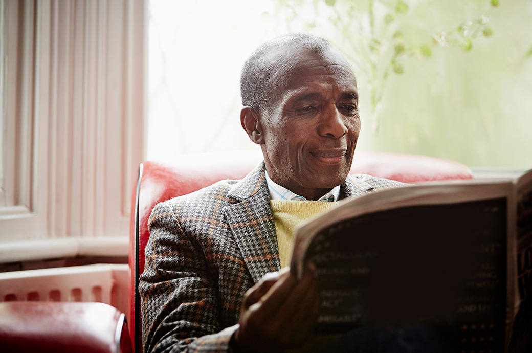 starší holohlavý muž tmavé pleti sedí v červeném křesle, čte si noviny a na tváři má lehký úsměv