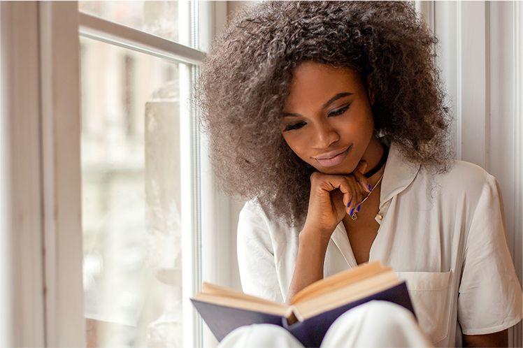 mladá žena tmavé pleti s kudrnatými vlasy v bílém tričku sedí u okna, čte si knížku a rukou si podpírá bradu